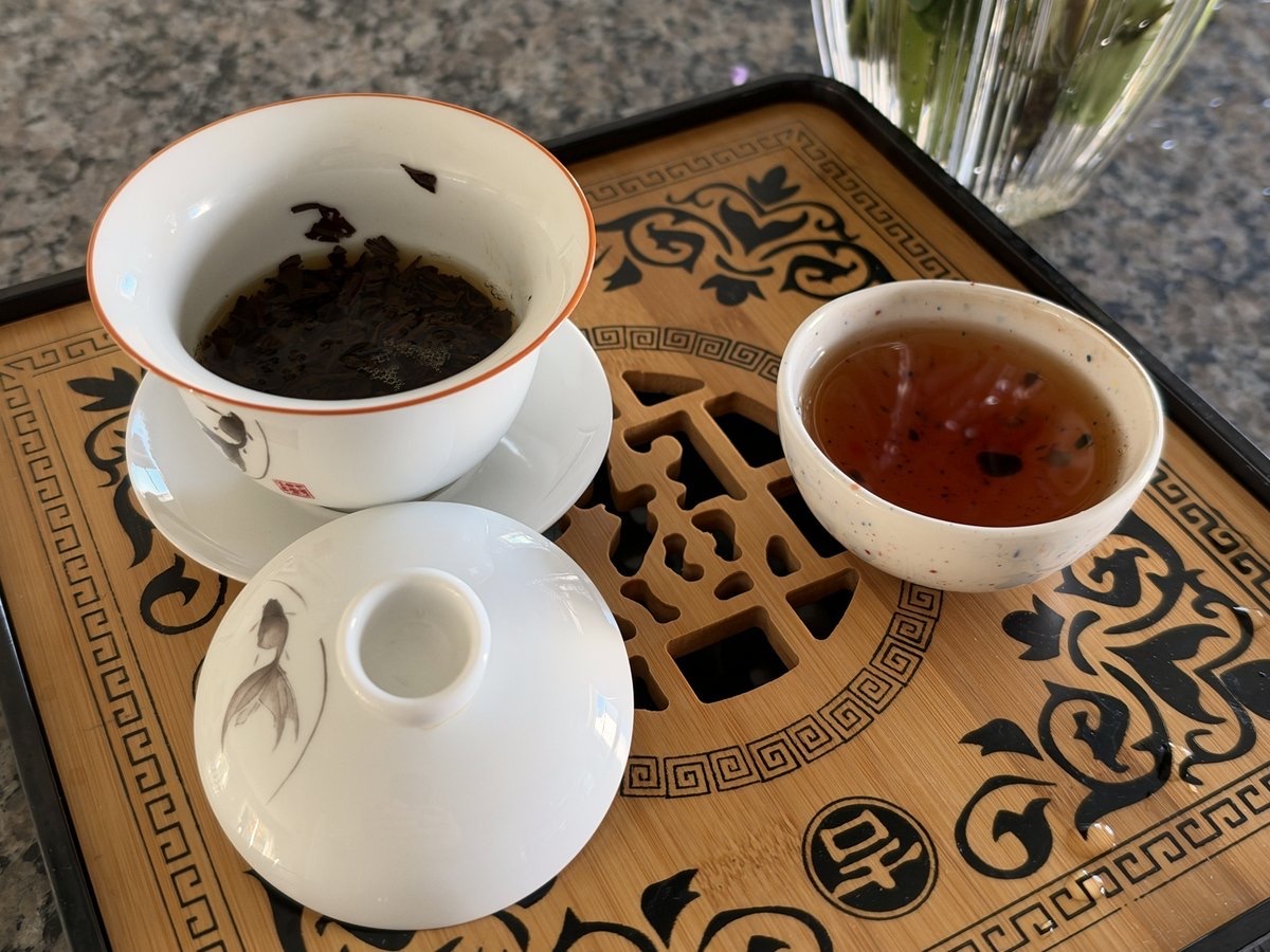 A white gaiwan of steeped leaf and a small cup of amber tea on a carved wooden tray